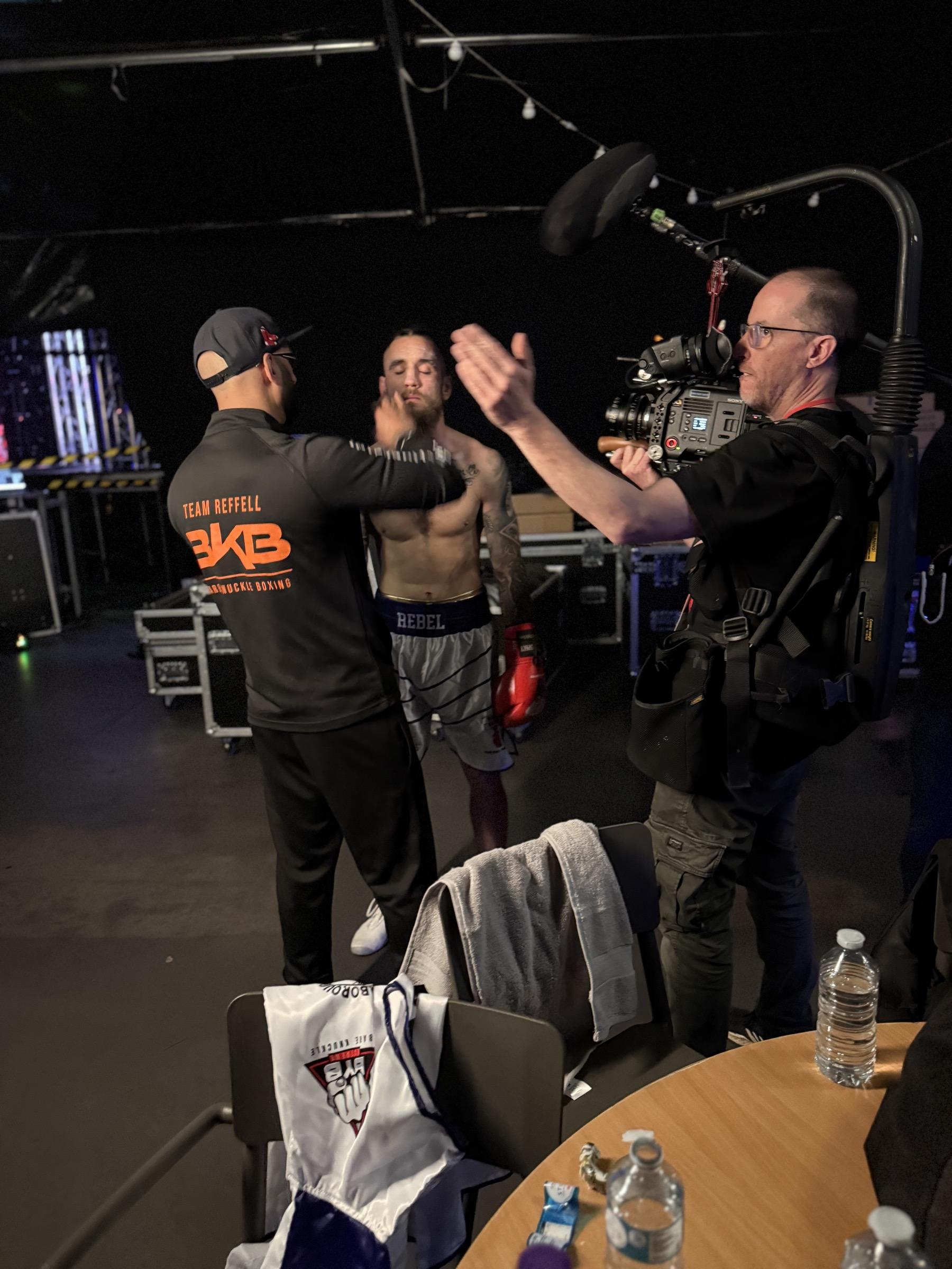 A camera operator films backstage as a cornerman wraps the hands of a shirtless, tattooed boxer wearing red gloves and branded shorts.