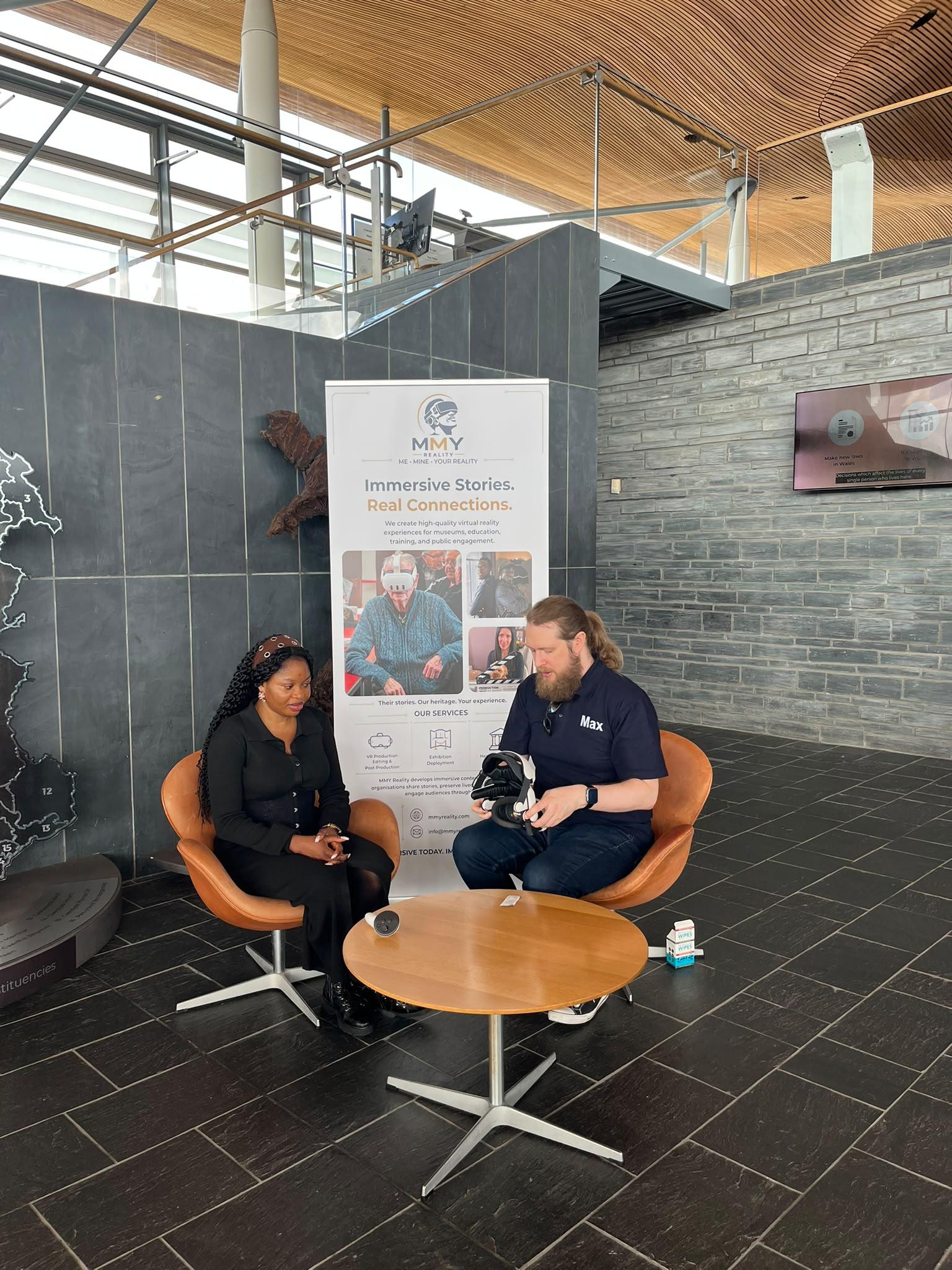 Two people sit in orange armchairs in a wood-and-slate hall, one holding a VR headset, beside an MMY Reality pull-up banner reading 'Immersive Stories. Real Connections.'