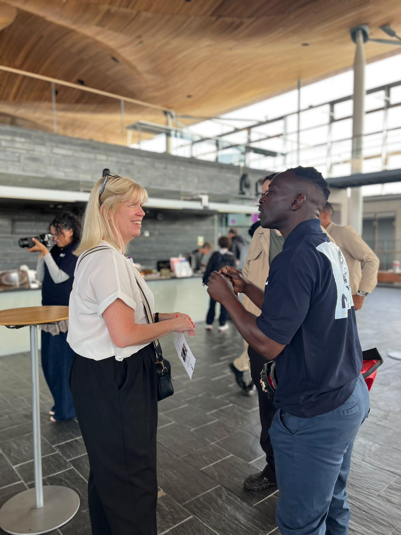 A blonde woman and a man in a navy polo shirt talk in a wood-ceilinged, slate-walled hall, with other attendees and a photographer in the background.